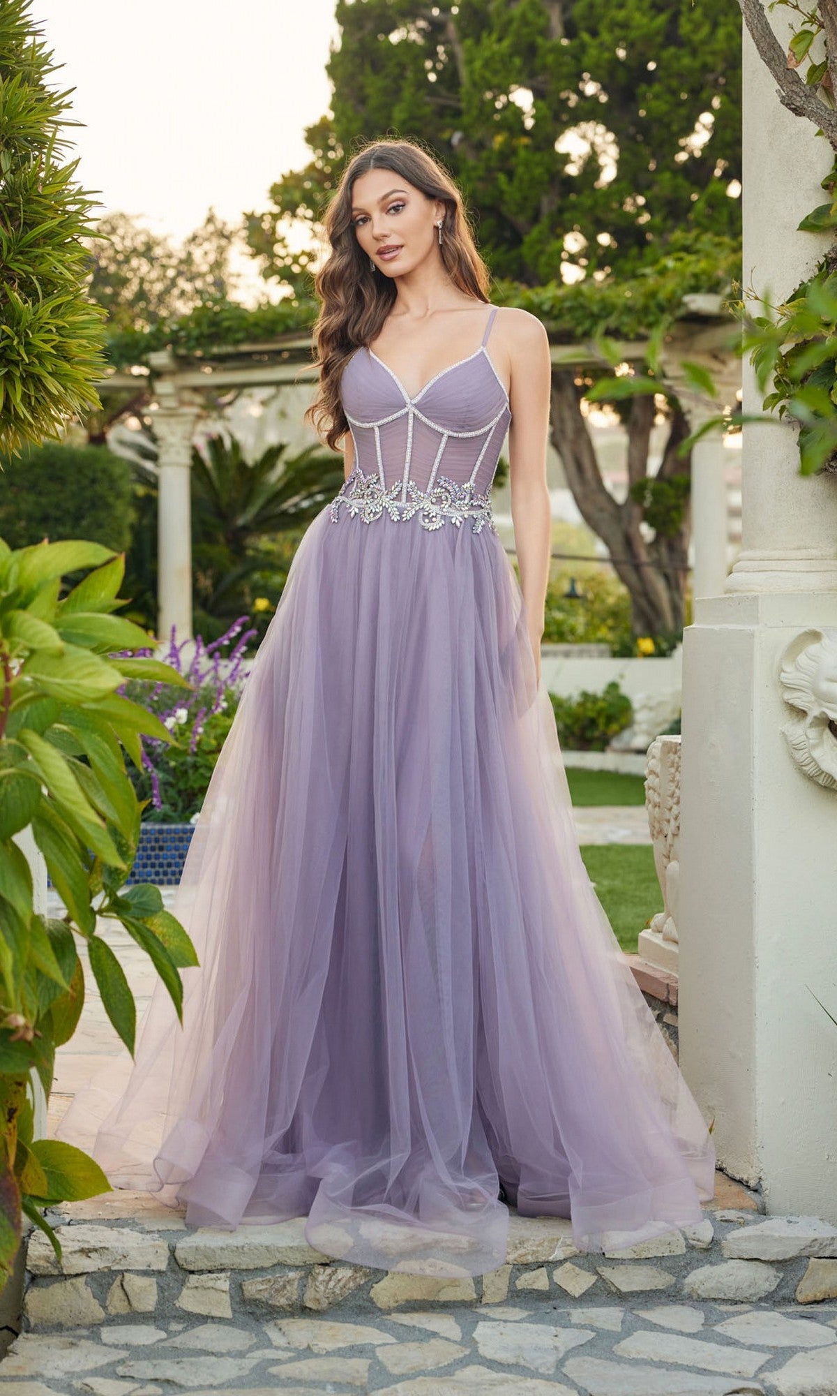 A woman stands outdoors surrounded by greenery and stonework, wearing the Long Prom Dress: Juliet JT2638G, featuring an a-line silhouette, light purple tulle skirt with floral embellishments, thin straps, and a structured bodice.